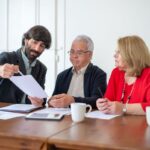 Trois individus analysent des papiers lors d'une réunion dans une pièce avec une table en bois