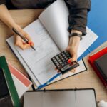 Person écrivant dans un livre avec une calculatrice, classeurs et téléphone sur une table