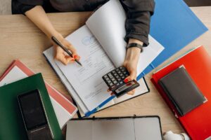 Person écrivant dans un livre avec une calculatrice, classeurs et téléphone sur une table