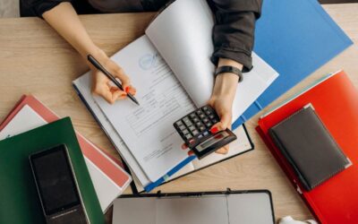 Person écrivant dans un livre avec une calculatrice, classeurs et téléphone sur une table