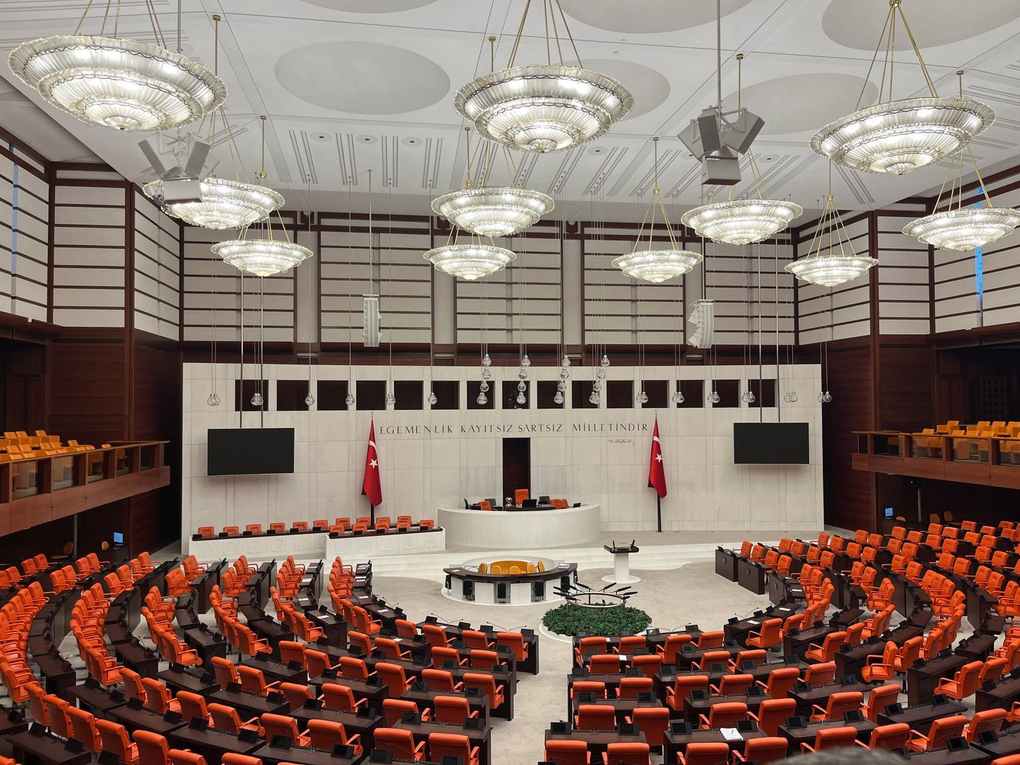Salle de conférence avec amphithéâtre, sièges rouges, bureau central, drapeaux, écrans, éclairage suspendu