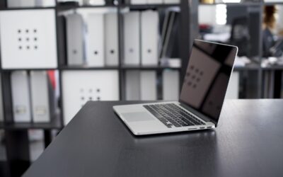 Un ordinateur portable ouvert sur un bureau, classeurs derrière